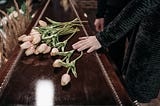 A woman’s hand lays gently on the lid of a polished casket. It’s dark brown in colour, and pale pink tulips are left by the mourners on the lid.