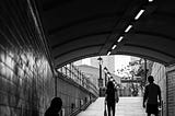 A black and white photo of a homeless person sitting under a bridge with two people walking by.