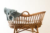 A woven bassinet/moses basket with white bedding and a grey knit baby jacket draped on one end.