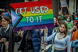 A Pride parade with several people marching. The two people in the foreground are holding a large Progress Pride flag on which is written in block letters, “GET USED TO IT.”