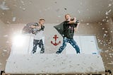 Tow boys pillow-fighting on a bed with feathers flying.