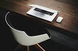 A Macbook with a magic mouse on a wood table next to a futuristic chair