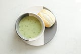 A bowl filled with creamy, parsley soup with a crusty roll on plate