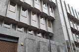 A grey marble clad facade with each of four floors cantilvered out beyond the floor below. Regularly spaced portrait shaped windows stand proud of the facade adding further to the blocky overall impression