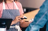 A person with an apron stands behind a checkout screen and scans an item for a person in a denim jacket.