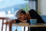 Photograph of a young woman in a beret waiting in a coffee shop, resting her head on her arm.