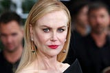 Foto da atriz Nicole Kidman. Nicole é uma mulher beem branca, com cabelos loiros lisos e compridos. Está com batom vermelho nos lábios e encara a câmera numa expressão séria. É possível ver parte da sua roupa preta.