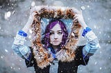 Young girl with purple hair and serious face pulling up her head covering to shelter from the rain and snow