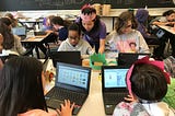 Classroom of students working on Scratch using personal computers. A teacher in pink crown assists students in the upper left side of the image.