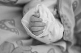 Black and white photo with closeup of an infant’s hand in an oversized sleeve. The baby’s sleeve with hand is surrounded by a blanket with non discernable pattern in gray tones.