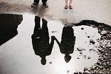 A couple reflected upside down in a puddle of water, holding hands.