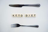 Depicted is a knife and fork laid horizontally on a white background. In the middle are white tiles spelling “KETO DIET”.