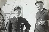 Phot depicts two naval captains standing outside in the sun, in their uniforms, laughing and one is smoking.