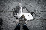 A person gazing into a puddle of water in cracked cement, the puddle reflecting an airplane and skyscrapers.