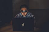 Image of a person sitting behind a laptop in the dark with sunglasses on