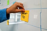 A person places a sticky note on the wall with the words ‘HOW TO’, signaling a focus on instructions, guidance, or problem-solving.