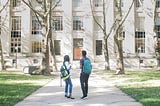 A female and male student with rucksacks on their backs standing in front of a college building