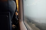 A person sitting inside a train gazing outside the window at the foggy fields.