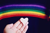 A person holding a pink heart with a cloth rainbow in the background.