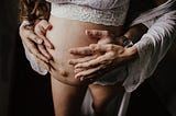 The doctor is in, and he’s bred me. Pregnant erotica. Doctor breeding patient erotica. A man stand behind a woman with their hand on her pregnant belly. The pregnant woman is wearing white lace.