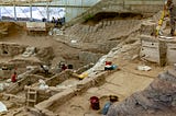 Photograph of an archaeological excavation in progress, showing exposed stretches of stone buildings and other features at different depths, with archaeologists digging or surveying separate portions of the site.