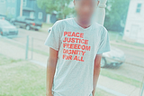 A young man with a blurred face wears a t-shirt that says “Peace, Justice, Freedom, Dignity for All.”