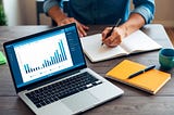 A laptop open on a desk with a graph of cryptocurrency prices, a notebook, and a pen. A person is looking at online tutorials on how to start crypto trading