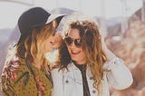 Two women one wearing a hat and the other leaning in and smiling to hear a secret.