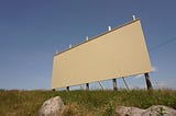 A blank, white billboard on the side of a deserted highway with some dry rocks and sun damaged grass.