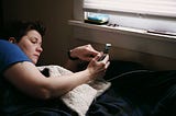 A woman is lying in bed, scrolling through her phone. The surroundings are barren, with just a few items on a windowsill.