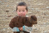 A small Asian child carried a brown lamb or baby goat in their arms, looking straight at the camera. against a plane dirt background.