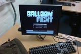 A computer screen on a desk with keyboard and mouse showing the title screen of the game “Balloon Fight” in black and white.