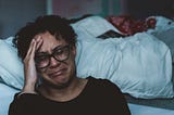 A woman, seated on the floor beside a bed, cries in anguish