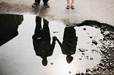 a couple holding hands looking at their reflection in a puddle.