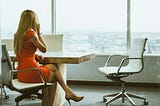 Woman in a red dress sitting at a conference table on the phone. The photo is meant to depict a female leader as the article is about women in leadership sometimes upholding patriarchal, misogynistic standards.