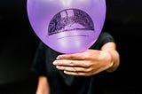 Man in black t-shirt holding a purple balloon in front of his face with a hand-sketched sad face on the balloon.