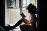 A young black women sitting by the window reading a book.