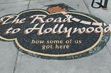 Sign on the sidewalk that says, “The Road to Hollywood: how some of us got here.”
