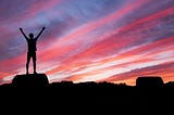 The dark figure of a man standing on rocks holding his arms high into a stunning red, orange and purple sky.