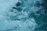 Turbulent sea from above — with some parts bluer, some whiter, it’s unclear what is going on but there are whirls and waves and foam