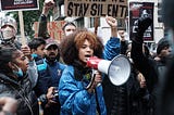 A femme presenting, POC activist with a megaphone standing up for justice with supporters surrounding her holding protest posters supporting socialism.