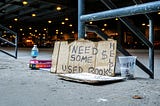 A sign reads, “Need some money, used books,” next to a cup for change.