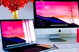 A Macbook and an iMac on a desk with red flowers.