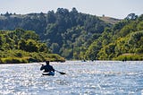 Photo of a man in a kayak paddling in water. Green hills are in the distance.