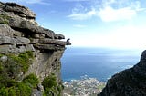 A mountain climber is sitting at the edge, overlooking a small coastal village.