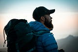 A young, bearded hiker with backpack stares into the distance