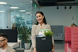 Happy young lady former employee is quitting job leaving office room with box of things walking and talking to ex-colleagues. People and employment concept.