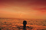 silhouette of a boy’s head above the water as he overlooks an orange sunset