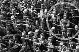 Man standing with his arms folded at Nazi rally where everyone else is saluting