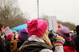 A blonde woman wearing a pink hat drinking a coffee, at the women’s march.